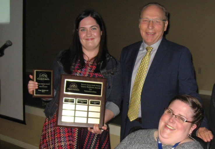 Paige McDonnell holding her award to show the camera. She has straight dark-brown hair and wears a bright patterned dress. She is standing with an older gentleman and a younger woman is a wheelchair.
